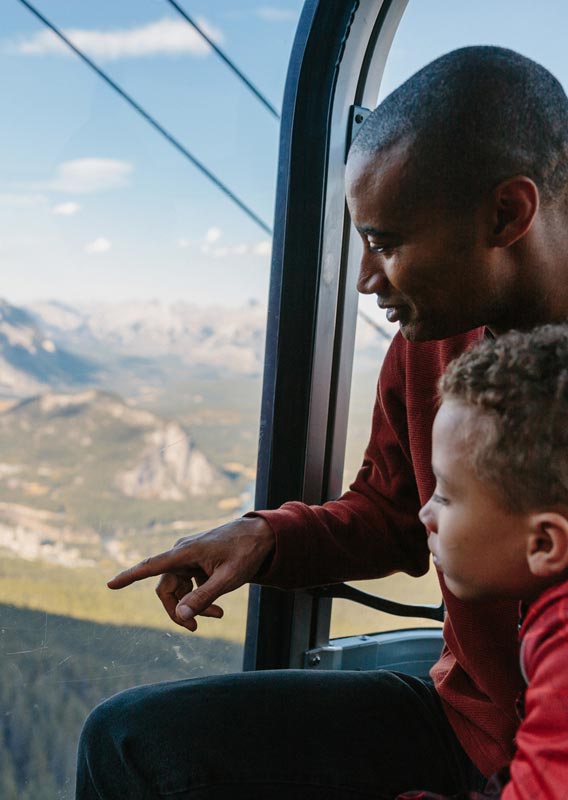 A father and son look out of the Banff Gondola. The boy points out the window with excitement