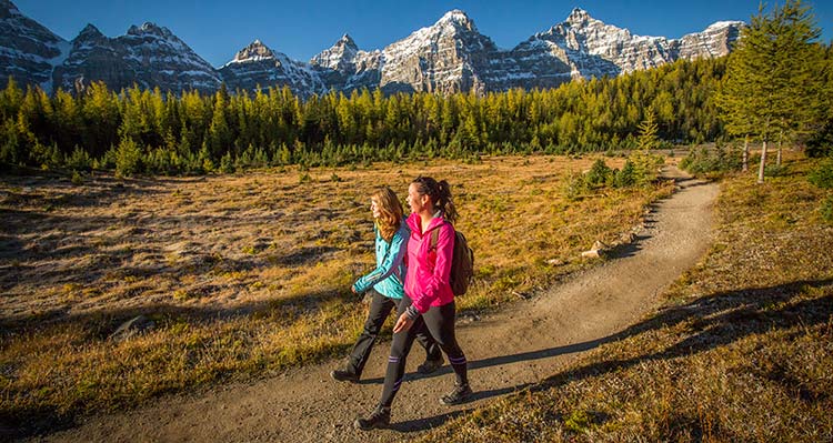 Two hikers walk on a meadow path below tall mountains.