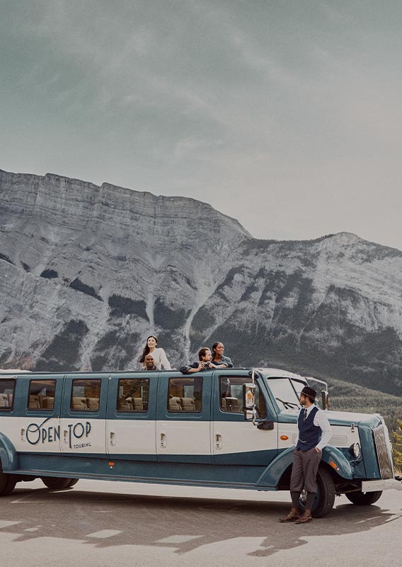 A driver stands out front of a historic-style bus with a group of people looking out from the roof.