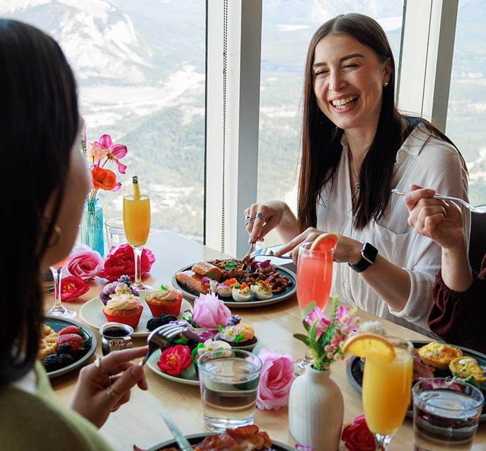 Three friends cheering mimosas at a window side table overlooking a valley.