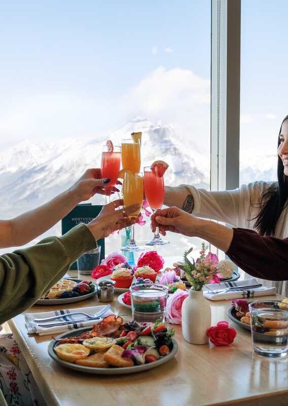 Four friends raise drinks in a cheers at a windowside table.