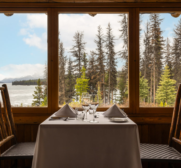 A dining table and chairs by the window, looking out to the trees and water, at the Maligne Lake Historic Chalet.
