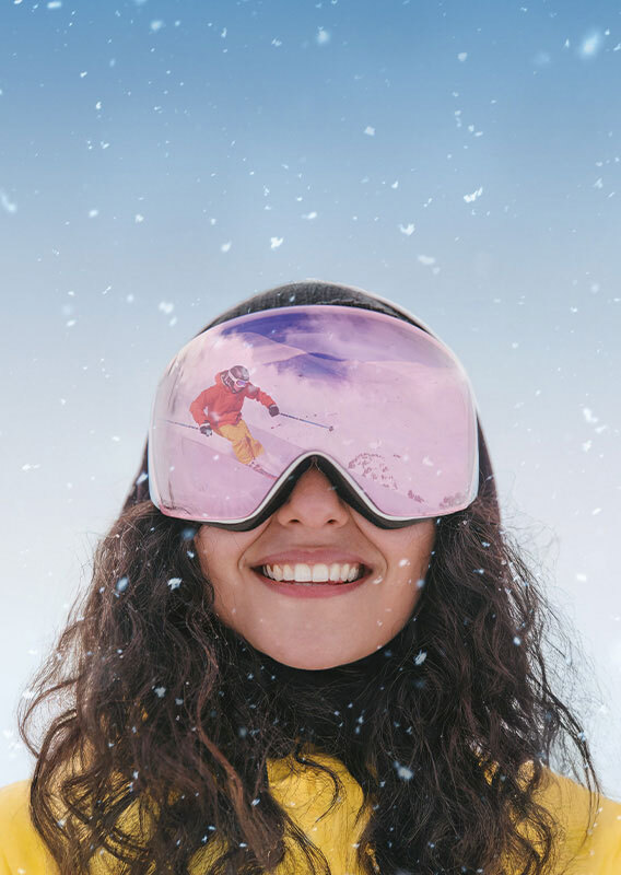 A woman smiles and a skiing scene is reflected in her goggles.