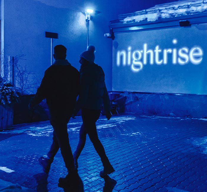 Two people walk past a wall with the word "nightrise" projected.