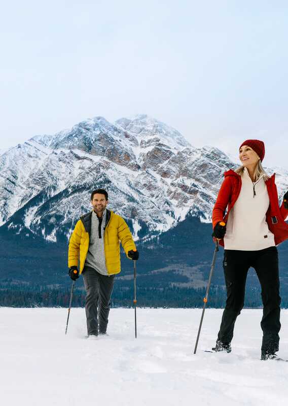 Two people cross country ski across a frozen lake below a tall mountain.