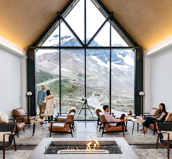 Small groups of people relax in a large lounge with a tall window looking out to a glacier.