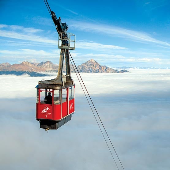 A red aerial tram ascends above a white cloud. Mountains are in the distance.