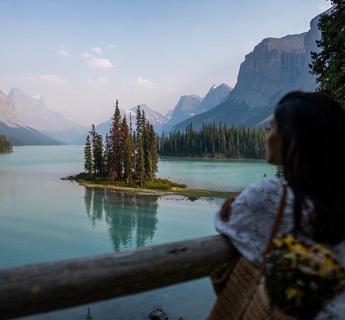A woman looks out from a lookout to a small peninsula on a lake below tall mountains.
