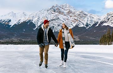 Two people skate on a frozen lake below tall mountains.